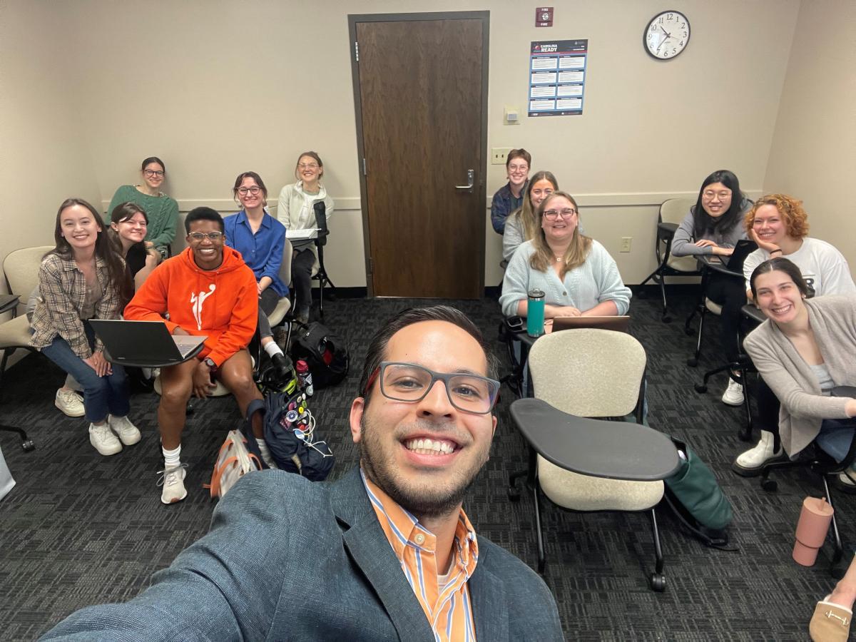 Man in glasses with a beard wearing an orange striped dress shirt and dark suit jacket takes a selfie with students standing behind him in a classroom.